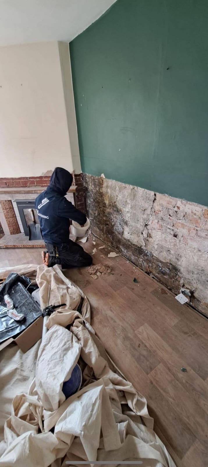 Callum on his knees hacking damp plaster off a wall beside a fireplace in a green-painted living room